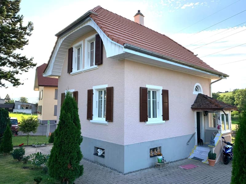 two-story house, light pink exterior, brown roof, several windows with shutters, small porch with statues and plant, driveway with parked motorcycle