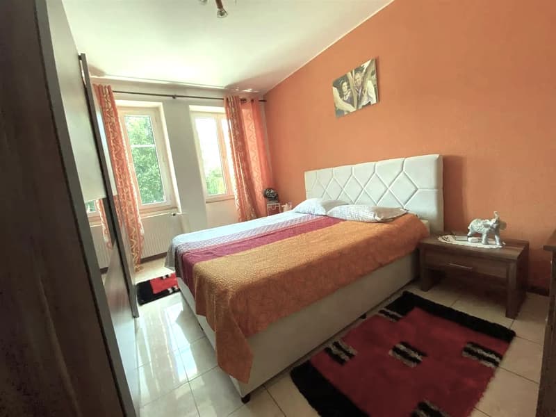 Double bed with two pillows, nightstand with figurine and books, orange curtains, beige wallpaper, beige tiled floor, two rugs, picture on the wall