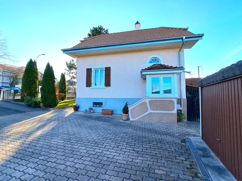 white facade with blue elements, brown roof, large driveway, shrubs and potted plants, garage with brown gate, street light, clear blue sky