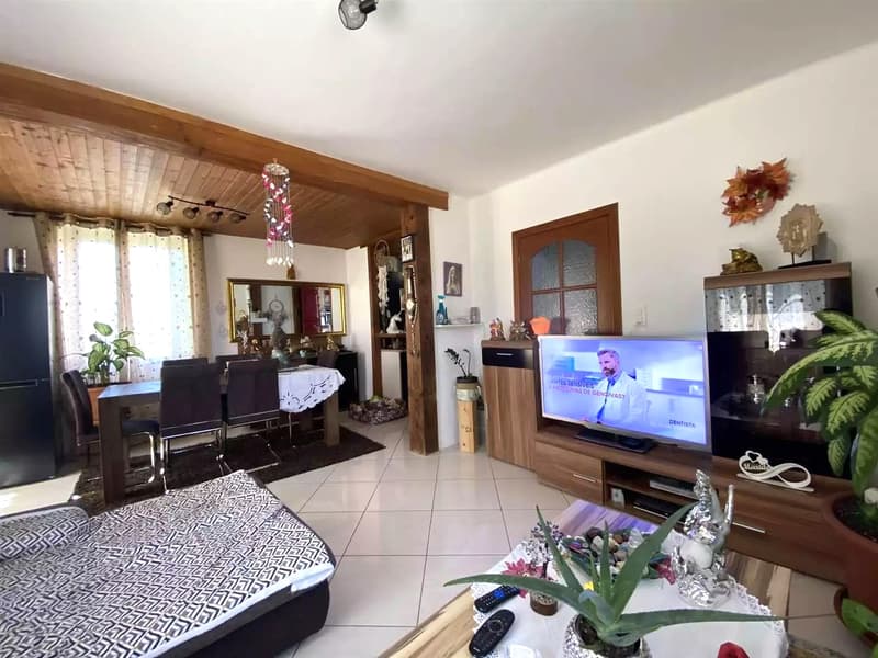 open floor plan, white tile floor, white walls, sofa, dining table, chairs, TV, plants, wooden ceiling, mirror, window