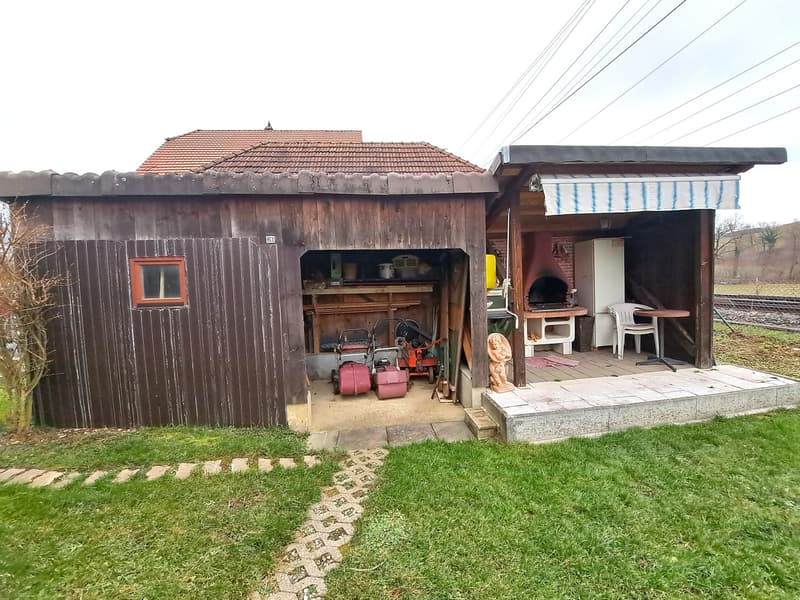 Garage with awning, attached to a house, backyard, small shed, lawn, electrical wires, trolley track, small trees, garden shed, outdoor table and chairs, outdoor table with a refrigerator, outdoor oven