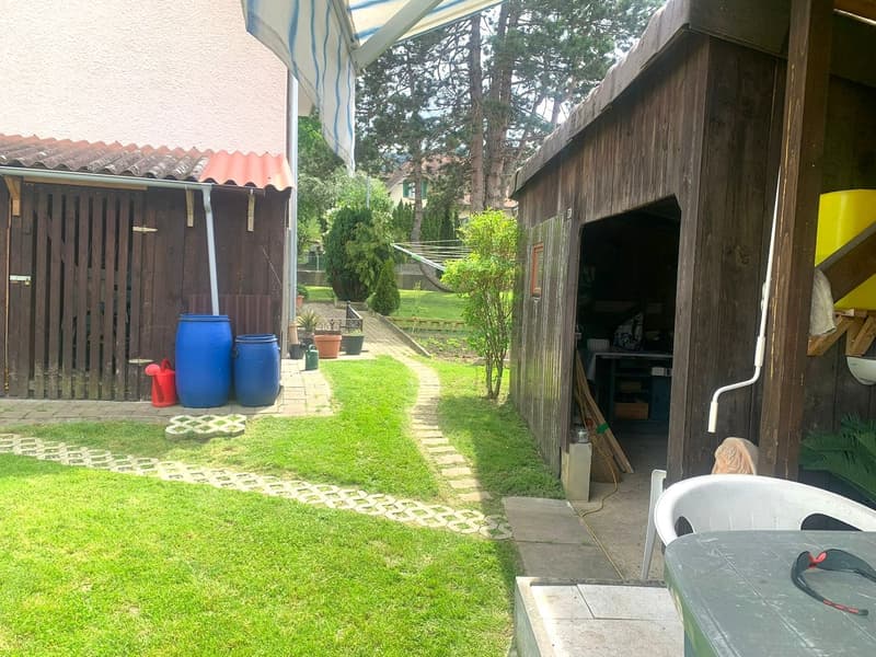 Wooden buildings, yard with garden, path leading to gate, two blue barrels, red watering can, and green plants.