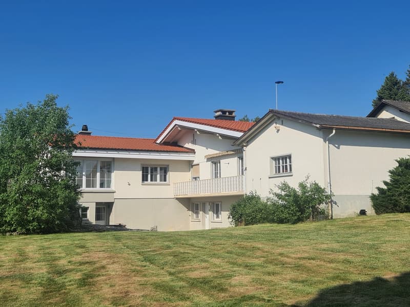 Two-story house with a red tile roof, white exterior walls, and large windows. The house has a balcony on the second floor. The property has a well-maintained lawn and is surrounded by trees.