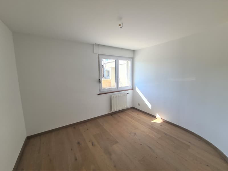 Spacious bedroom with white walls, hardwood flooring, and a large window providing natural light. The room appears to be empty and ready for furnishing.