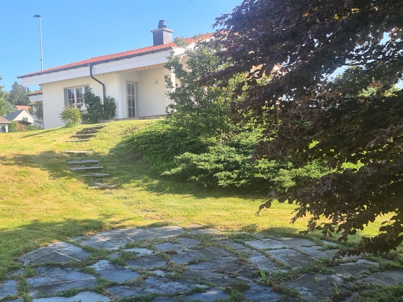 Two-story white house with red tile roof, surrounded by lush greenery and trees, with a stone pathway leading up to the house and a balcony or terrace visible on the upper level.