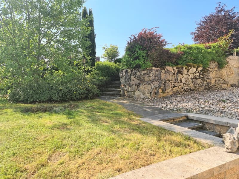 The image shows a well-maintained outdoor garden area with a stone pathway leading up to a set of stone steps. The garden is surrounded by lush greenery, including trees, shrubs, and a grassy lawn. There appears to be a small water feature or pond visible