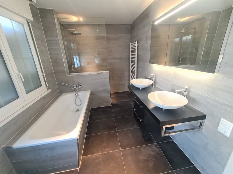 Large bathroom with gray tile walls and floors, freestanding bathtub, double vanity with vessel sinks, and a glass-enclosed shower area