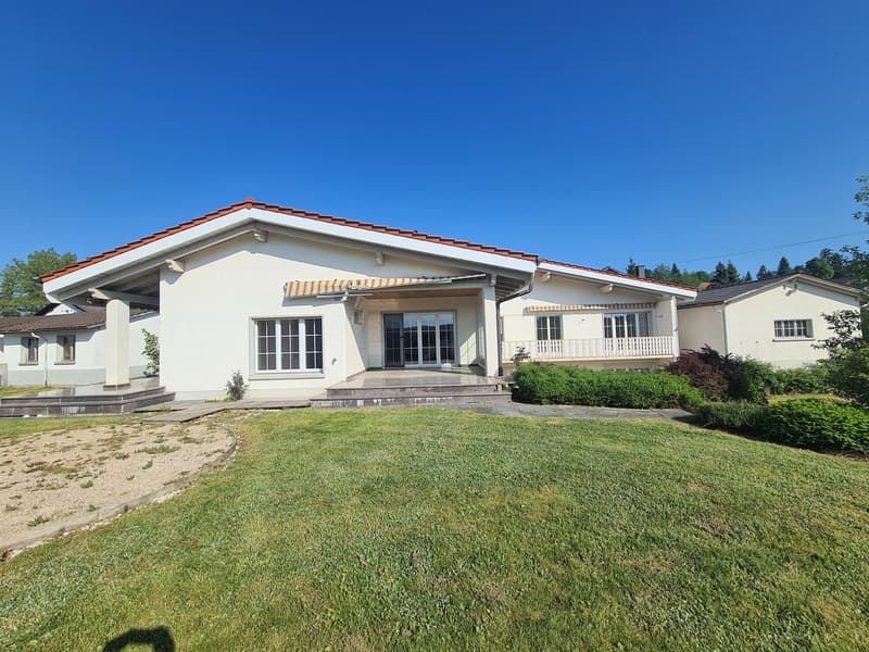 Single-story house with a red tile roof, white exterior walls, and large windows. The house has a balcony or terrace on the front. The property is surrounded by a well-maintained lawn and landscaping.