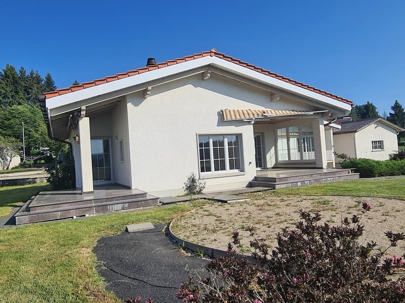 Single-story house with a red tile roof, stucco exterior, large windows, and a balcony or terrace. The house is surrounded by a well-maintained lawn and landscaping.