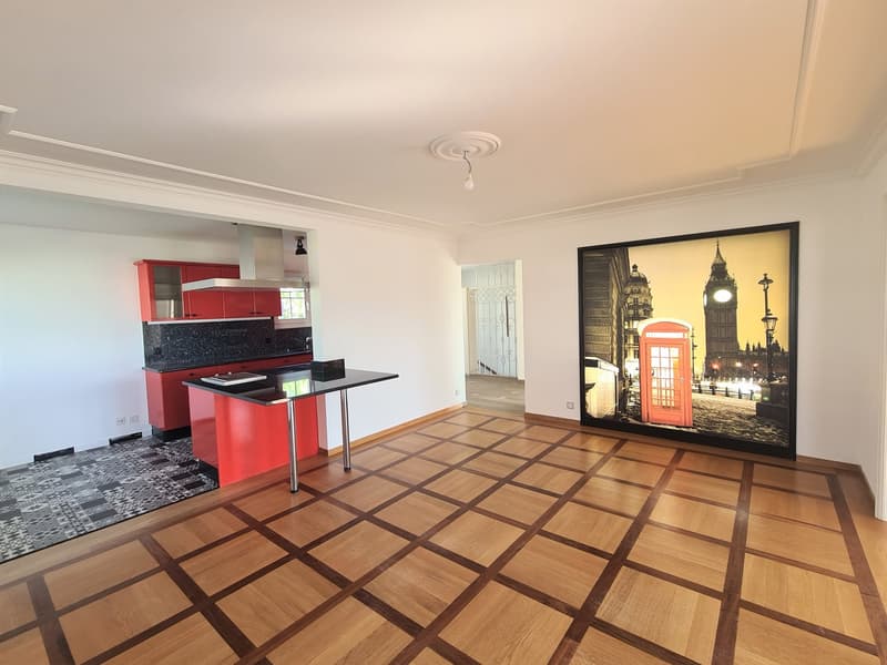 Open-concept living space with a modern kitchen featuring red cabinets and black countertops, a large framed artwork depicting a London street scene, and a patterned tile floor.