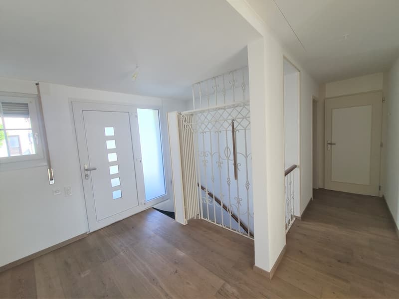 The image shows the interior of an apartment. The entrance features a white door with glass panels. The hallway has hardwood flooring and a decorative metal railing. The walls are painted in a light color, creating a bright and airy atmosphere. The space