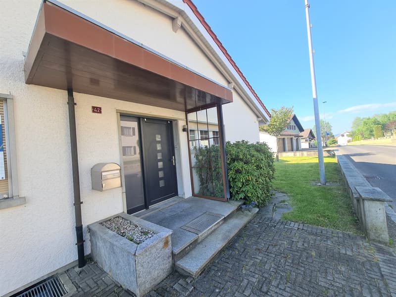 2-story residential building with a raised ground floor, brown roof, and a covered entrance with a glass door. The building has a small garden area in front with some plants and a mailbox.