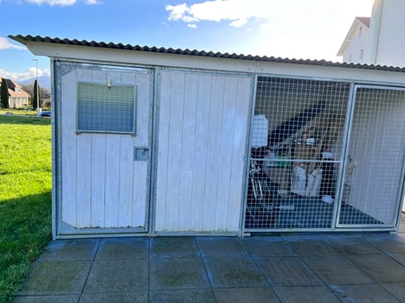 A small, single-story wooden shed with a corrugated metal roof. The shed has a garage-style door and a window. It is situated on a grassy area next to a residential property.
