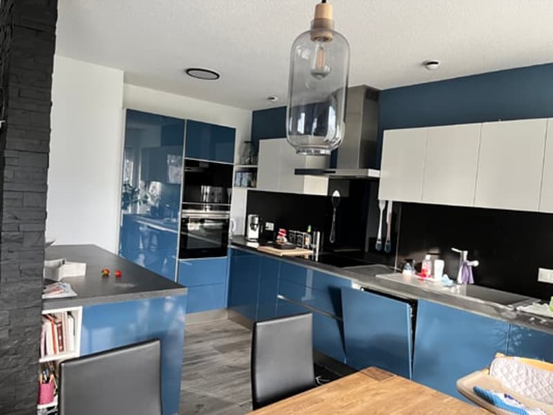 Modern kitchen with blue and white cabinets, black countertops, stainless steel appliances including a dishwasher, and a large pendant light fixture over a wooden dining table