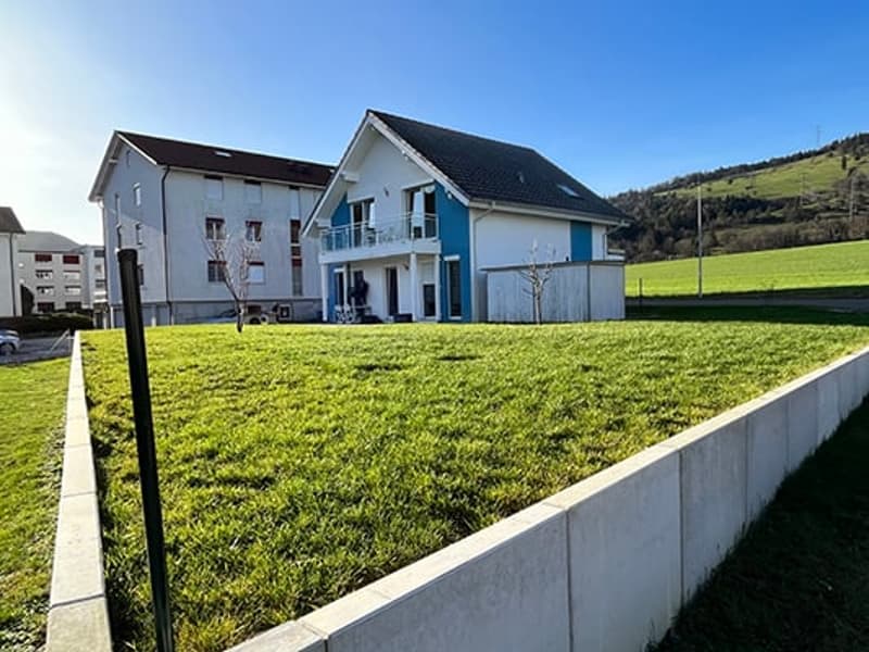 Two-story house with white exterior, black roof, and a garage. The house is surrounded by a grassy lawn and is situated in a rural or suburban area with hills in the background.