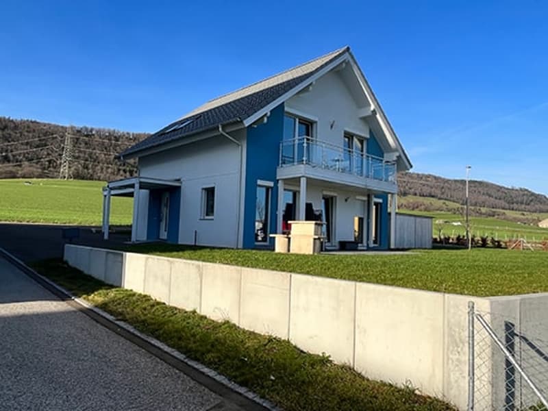 Two-story house with blue exterior, balcony, and surrounded by a grassy yard and hills in the background.