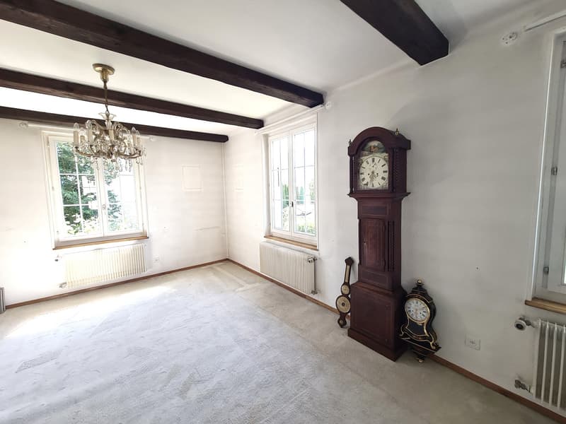 Spacious room with hardwood beams on the ceiling, large windows, a crystal chandelier, and an antique grandfather clock against the wall. The room has a neutral color palette and appears to be in a traditional or historic style.