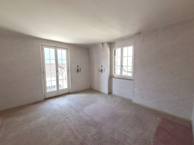 This image shows an empty room with white walls and a hardwood floor. The room has two large windows with white frames, and there are two wall sconces on the walls. The room appears to be in need of renovation or remodeling, as the floor and walls are unf