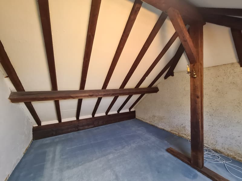 The image shows an attic space with exposed wooden beams and a concrete floor. The space appears to be unfinished, with a simple, utilitarian design.