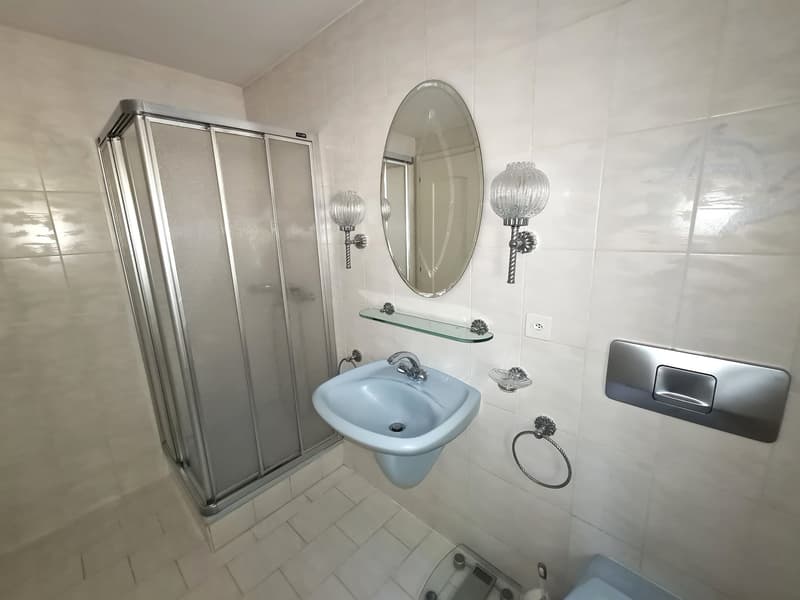 The image shows a bathroom with a glass shower enclosure, a blue sink, a round mirror, and decorative wall sconces. The bathroom has a tiled floor and walls.