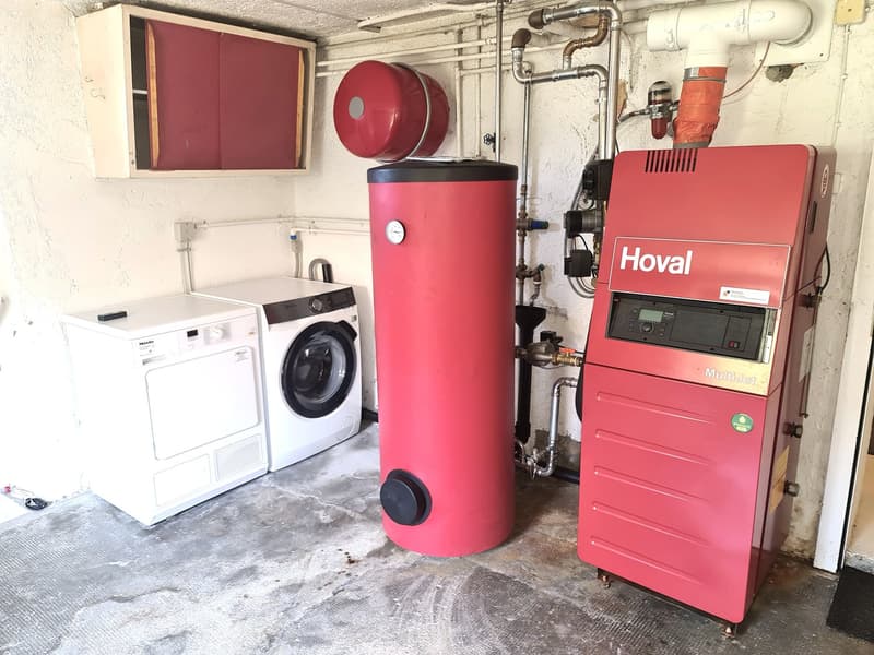 The image shows a utility room with a washing machine, a red Hoval boiler, and various plumbing and heating equipment. The room also contains a red storage cabinet and a red cylindrical tank, likely a water heater.