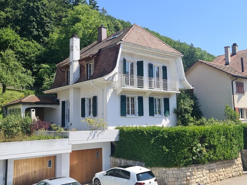 Two-story white house with a tiled roof, balcony, and garage. The house is surrounded by lush greenery and trees.