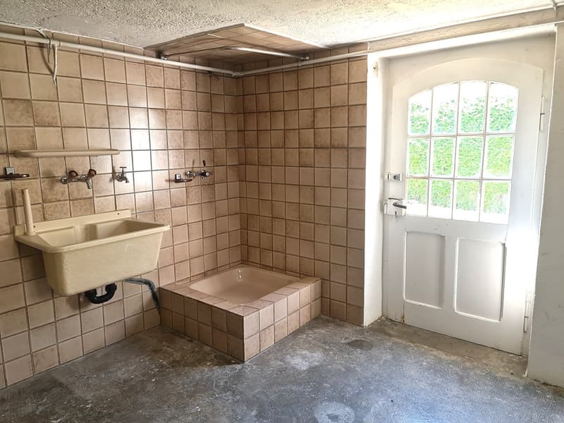 The image shows a bathroom with tiled walls, a sink, and a raised tub or shower area. The room has a window that provides natural light, and the floor appears to be concrete or a similar material.