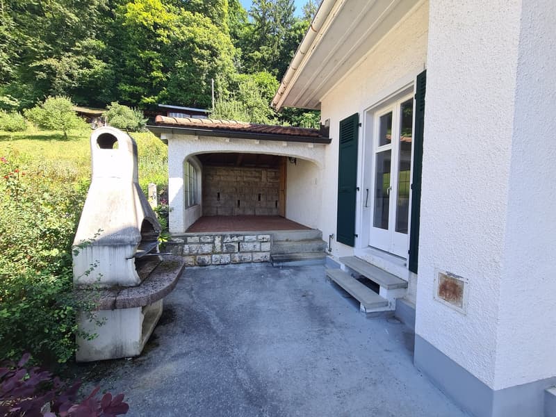 The image shows the exterior of a house with a covered entryway or garage area. The house has a white exterior with green shutters and a tiled roof. There is a stone pathway leading up to the entrance and the surrounding area is lush with greenery.