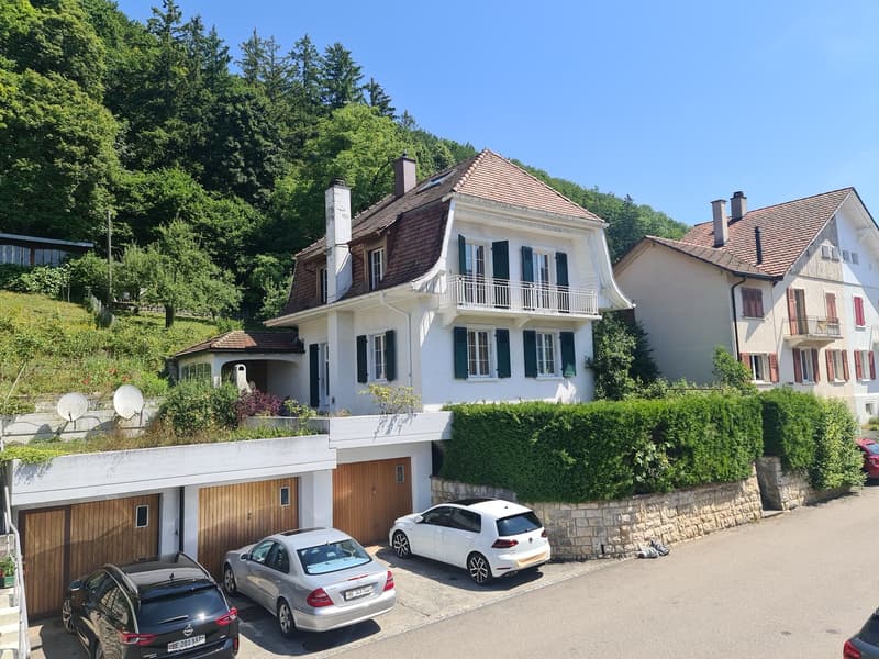 Two-story house with a red tile roof, white exterior walls, and black shutters. The house has a balcony on the second floor and a garage with parking spaces in front. The property is surrounded by lush greenery, including trees and hedges.