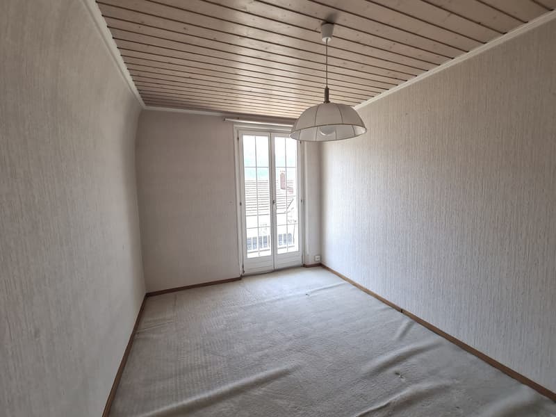 Empty room with white walls, wooden ceiling, and a sliding glass door leading to a balcony or terrace. The room has a pendant light fixture hanging from the ceiling.