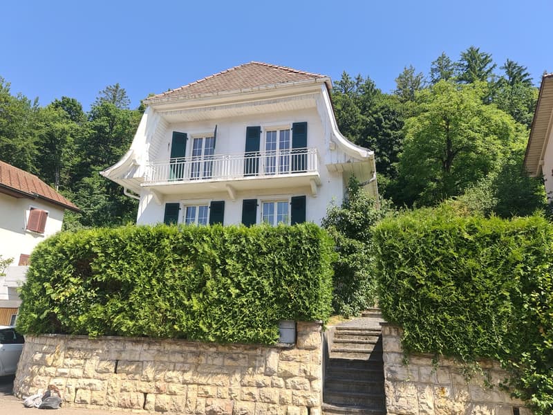 Two-story white house with a tiled roof, surrounded by lush greenery and trees. The house has a balcony on the second floor and stone steps leading up to the entrance.
