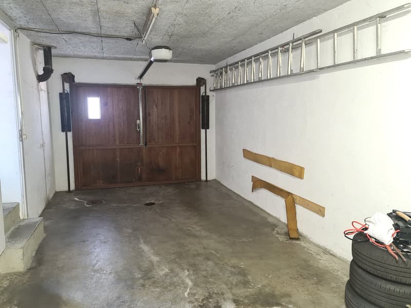 The image shows a garage space with a concrete floor, wooden paneled walls, and a window. There is a ceiling fan and some storage shelves along the walls. The garage appears to be used for storage, with some tires and other items visible on the floor.