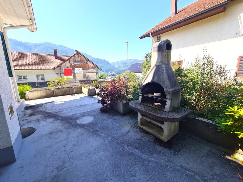 The image shows an outdoor area with a stone fireplace or oven, surrounded by plants and greenery. There are residential buildings visible in the background, suggesting this is part of a residential property or community. The area appears to be paved with