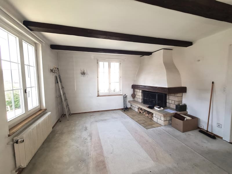 Large open living room with white walls, exposed wooden beams on the ceiling, a brick fireplace, and a ladder leaning against the wall. The room appears to be in the process of renovation or remodeling, with some boxes and items on the floor.