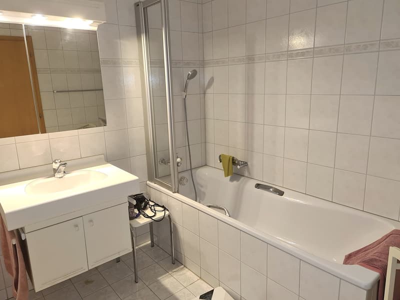 White tiled walls and floor, bathtub with shower, sink with vanity cabinet, large mirror, a towel, a chair in front of the bathtub.