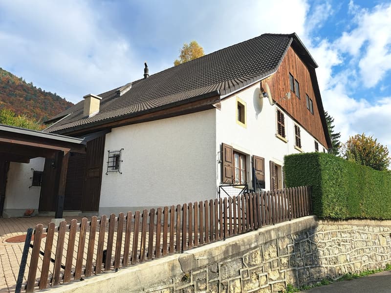 Traditional-style house with a slate roof, white and brown paint, wooden entrance, stone wall, garden, brick driveway, mountain in the background.