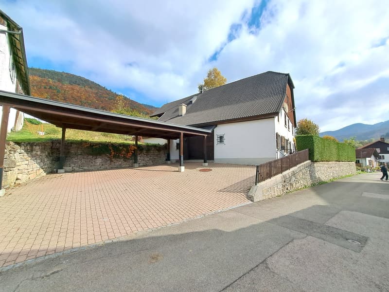 A house with a gray roof, white walls, brick driveway, stone wall, and plants. There's a covered carport, a grassy area with plants, and mountains in the background.
