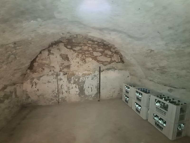 an empty, dim basement with stone arch and cracked concrete walls, water bottles stored in boxes