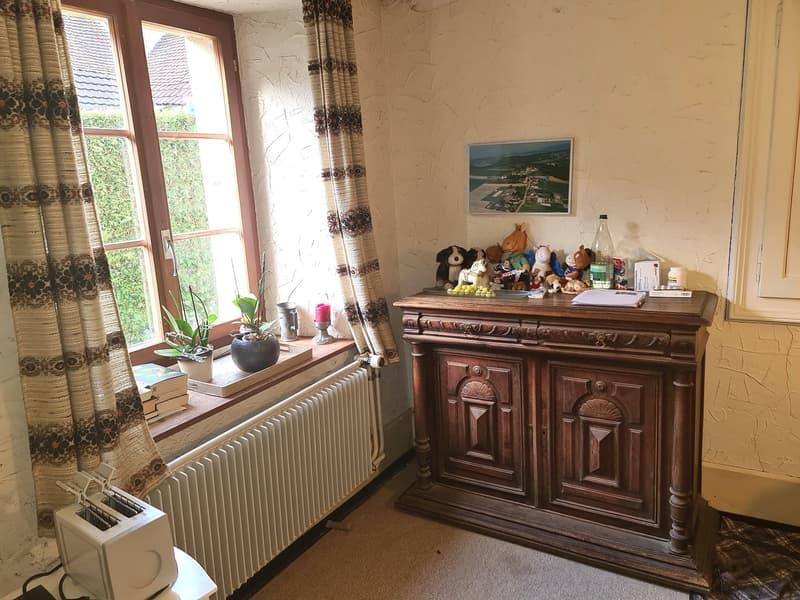 A room featuring a wooden cabinet, window with curtains, potted plants, picture frame, radiator, toaster, and various items on the cabinet.