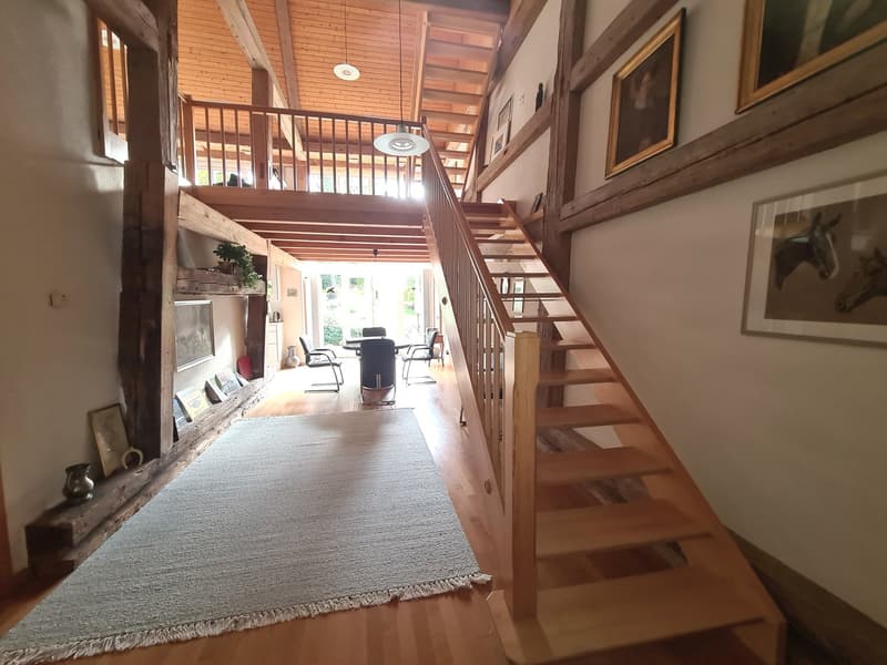 Entrance area, wooden stairs, wooden handrail, gray carpet, hardwood floor, framed pictures on the wall, wooden beams, balcony