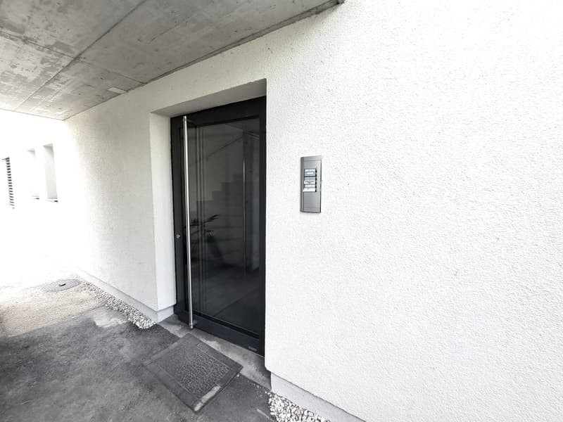 The image shows the entrance to a building. The entrance has a glass door with a metal frame. The wall surrounding the entrance is white and the floor is covered with small stones. There is a metal intercom system mounted on the wall next to the door.