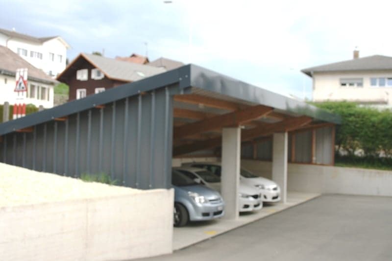 A multi-car garage or parking structure with a covered roof and open sides, located in a residential area with nearby houses and buildings.