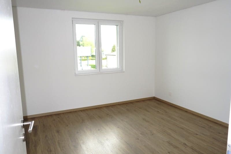 Spacious room with white walls, large windows, and hardwood flooring.