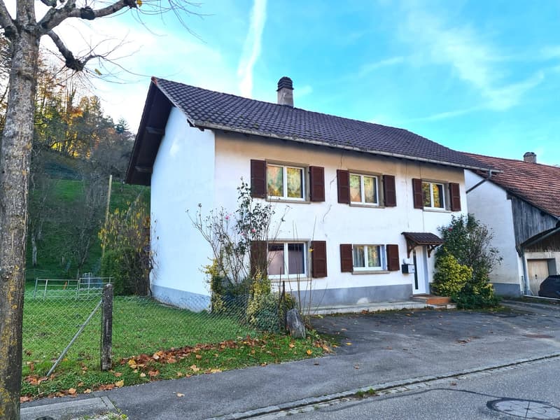 white exterior, brown tiled roof, raised ground floor, chimney, several windows, driveway, greenery around