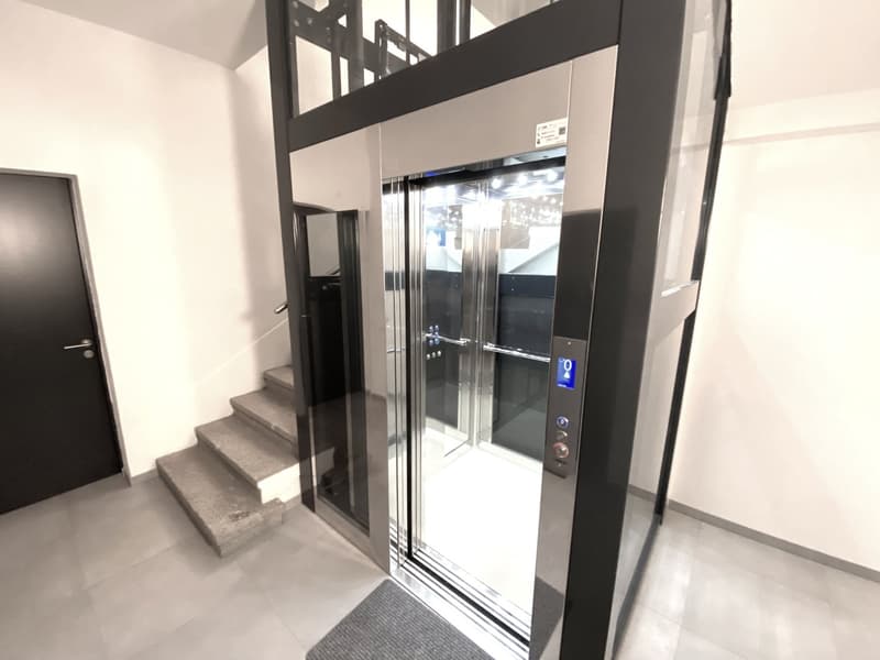 Modern elevator with glass doors and control panel, adjacent staircase, raised ground floor