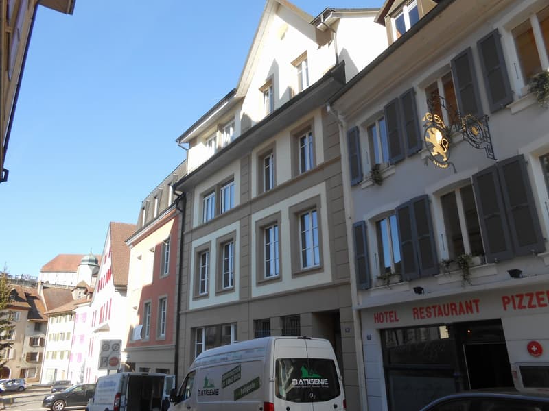 White and pink buildings with windows, some having shutters, a hotel and restaurant on the right, and cars parked on the street.