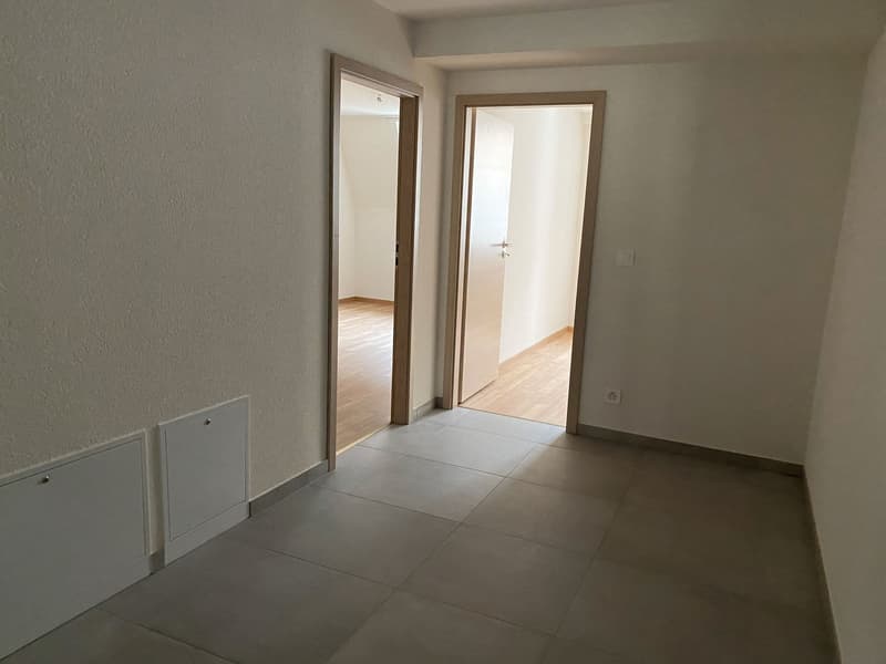Empty room with entrance hallway, wooden floor, white walls, and tiled flooring.