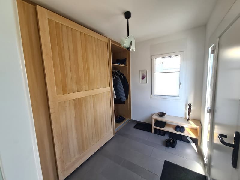 Large wooden wardrobe, clothes hanging on hangers, tiled floor, window, shoe shelves, decorative items, wall light