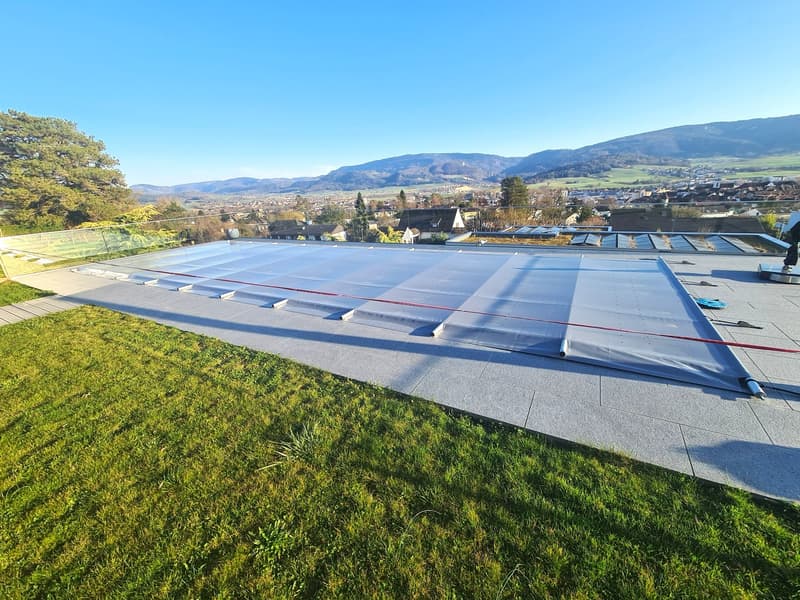 Rooftop area covered with a gray tarp, pathway with tiled flooring, lush green grass, glass railing along the edge, a distant view of buildings and mountains