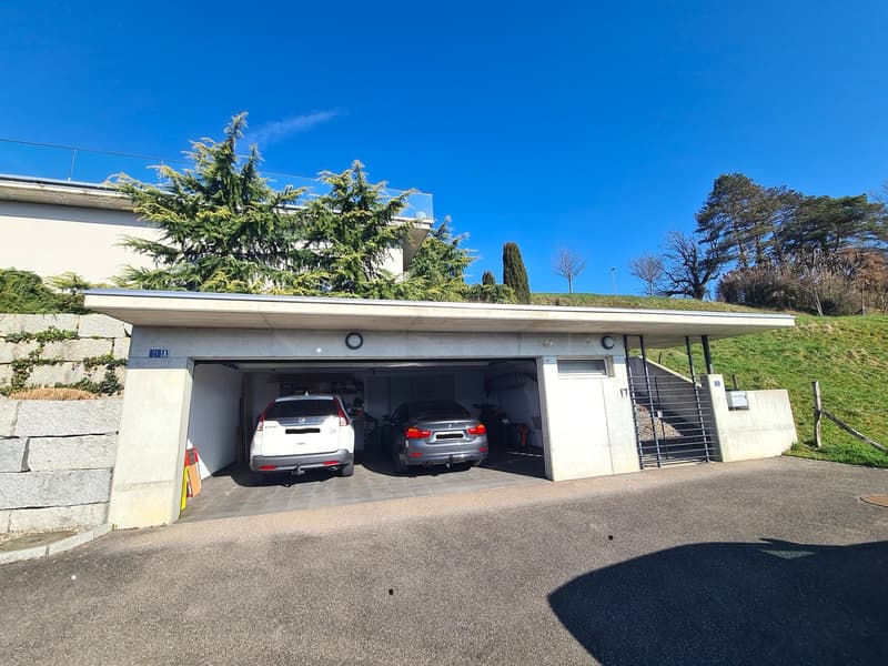Subterranean garage with two cars parked, white walls, concrete floor, glass door, stairs leading to the street.
